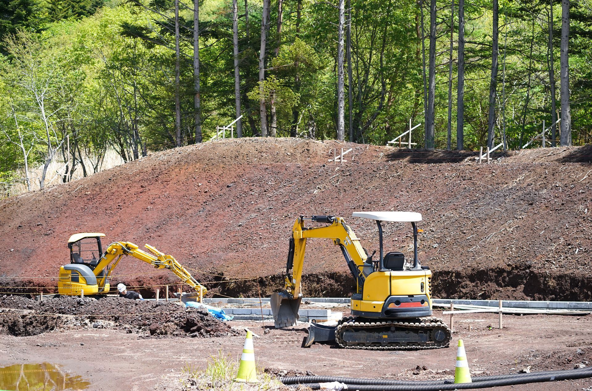 Two yellow excavators on a construction site, near a dirt mound and trees.