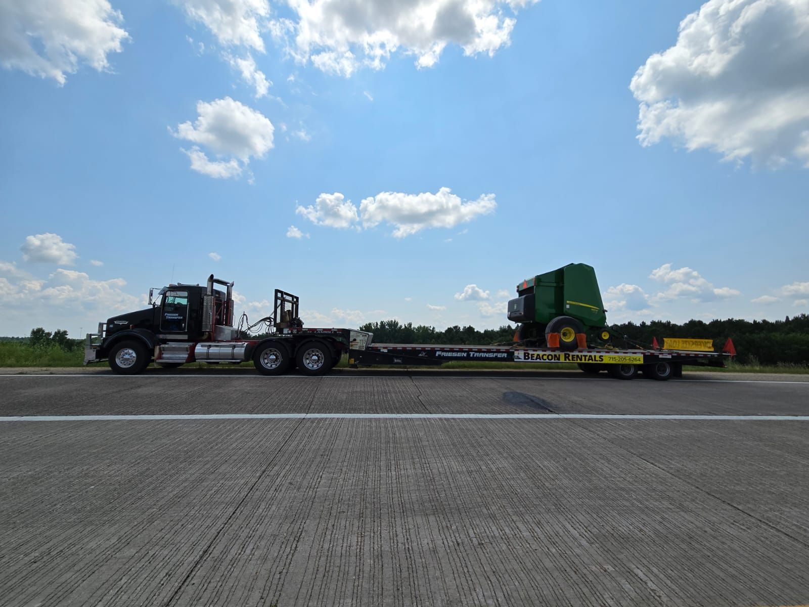 Black semi-truck hauling a green John Deere machine on a flatbed trailer on a highway under a blue sky.