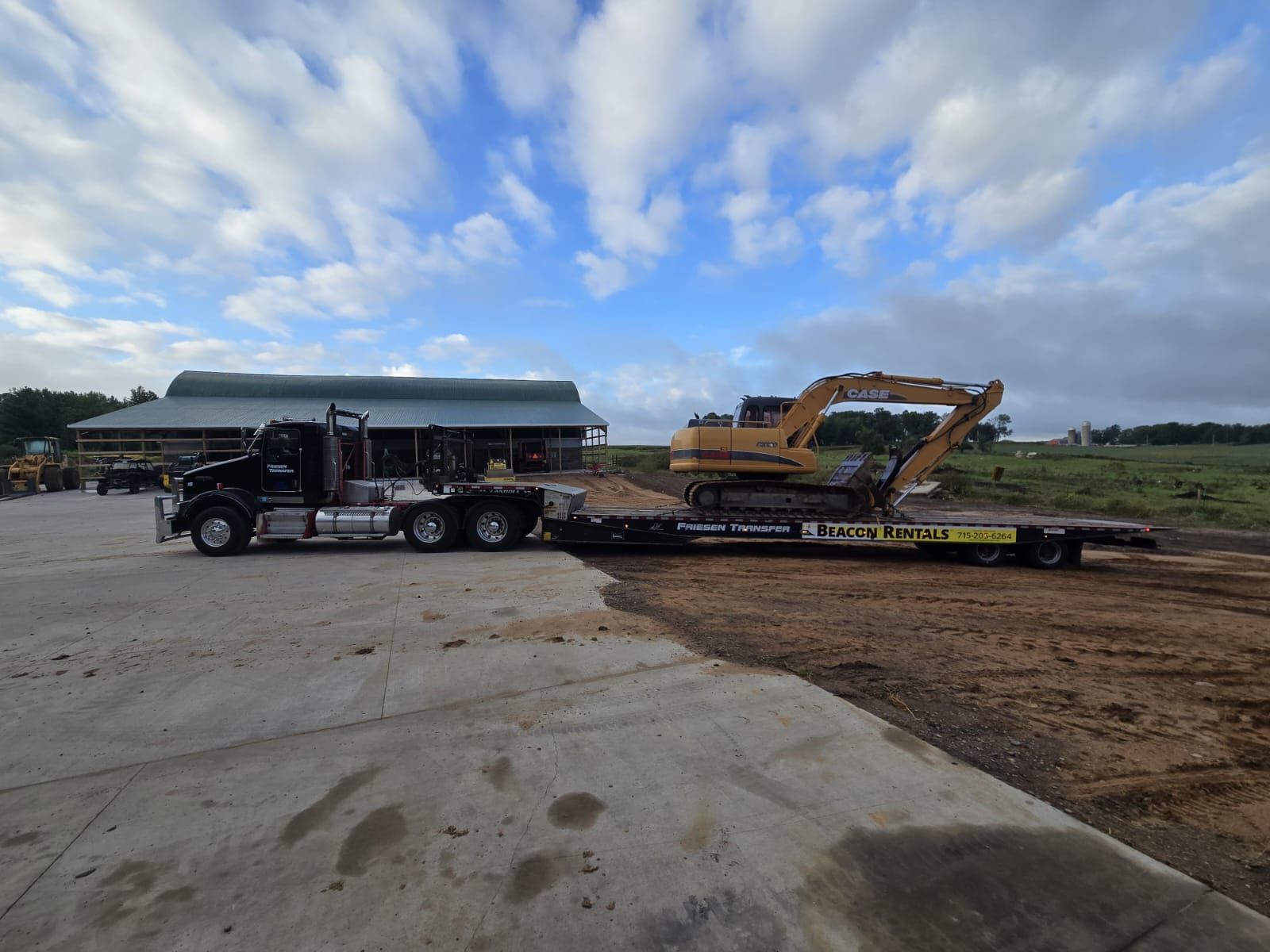 Black truck hauling yellow excavator on a flatbed trailer, parked on concrete in front of a building under a cloudy sky.