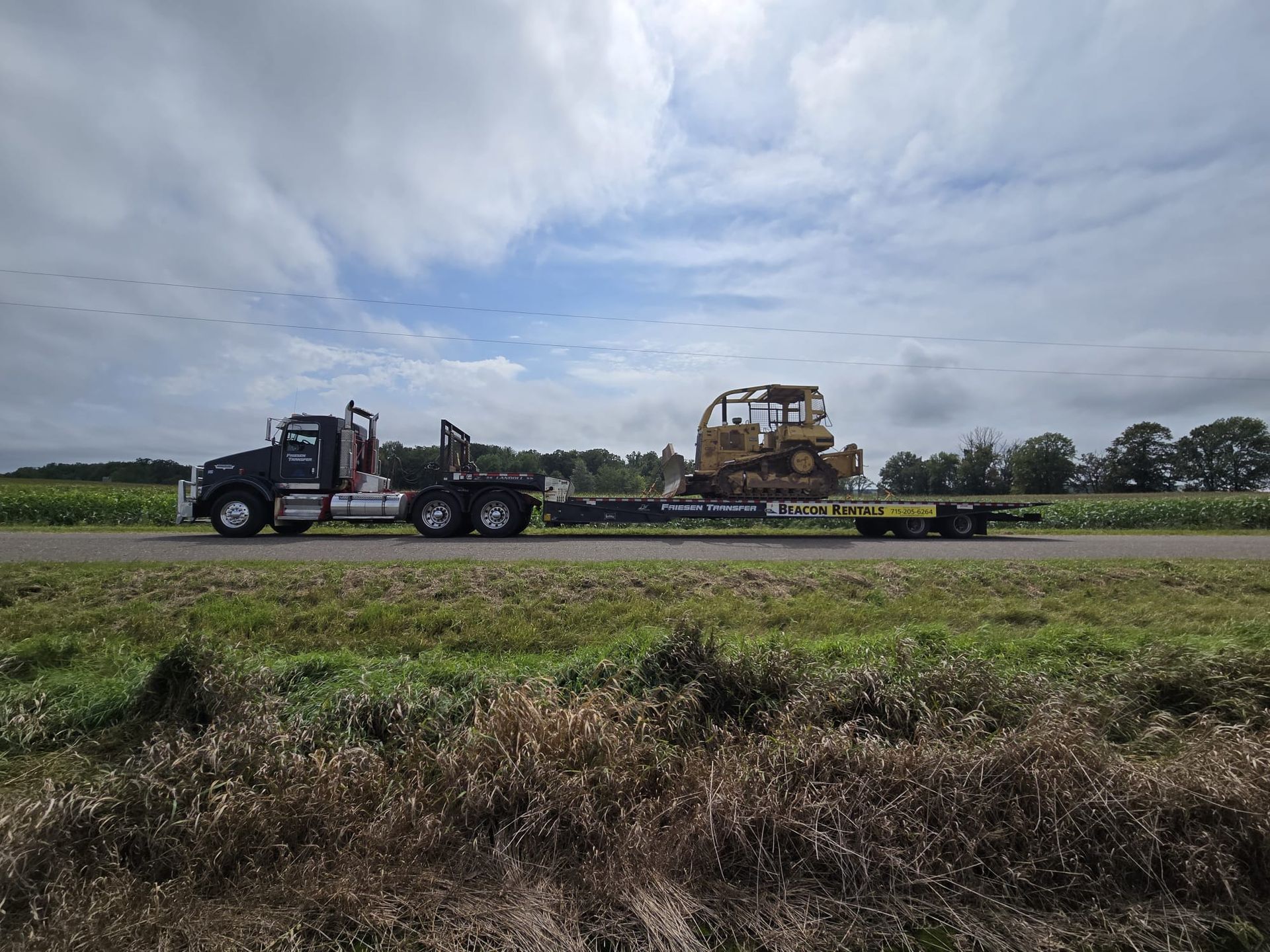 Black truck hauling a yellow bulldozer on a flatbed trailer, driving down a road, cloudy sky.