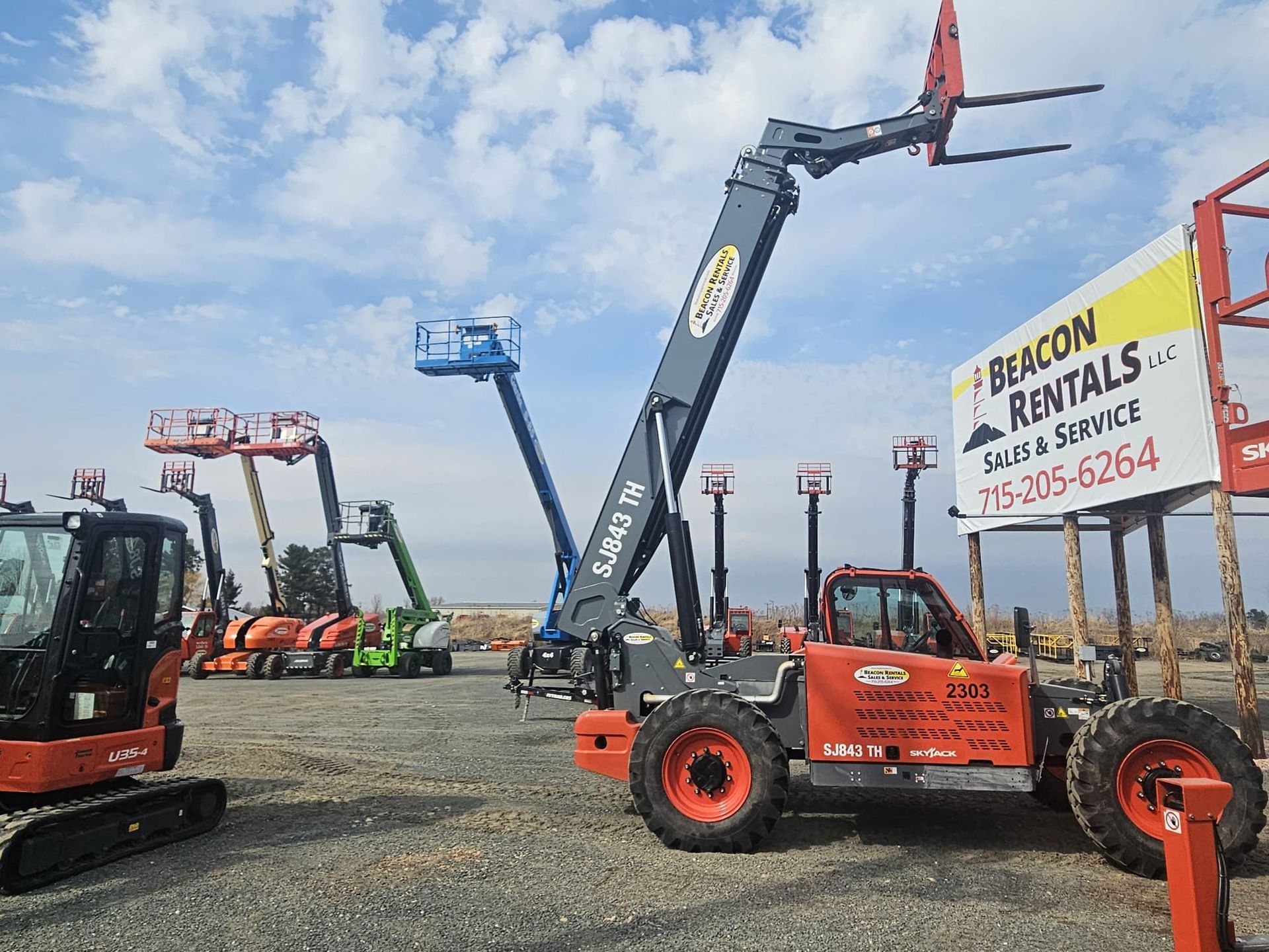 Orange telehandler with raised forks, in front of a rental business with other equipment.