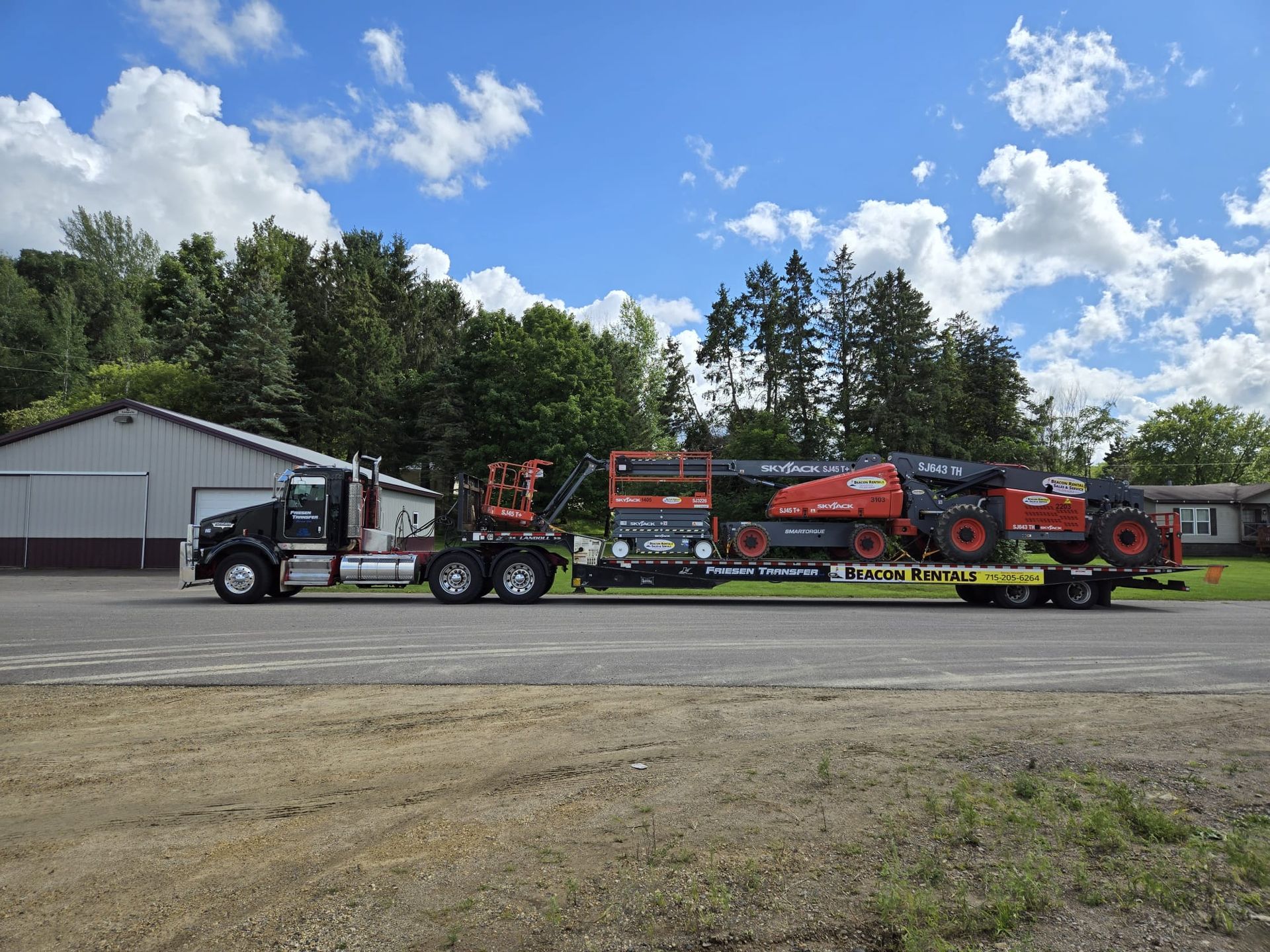 Black semi-truck hauling a trailer loaded with orange construction equipment on a gravel lot under a blue sky.