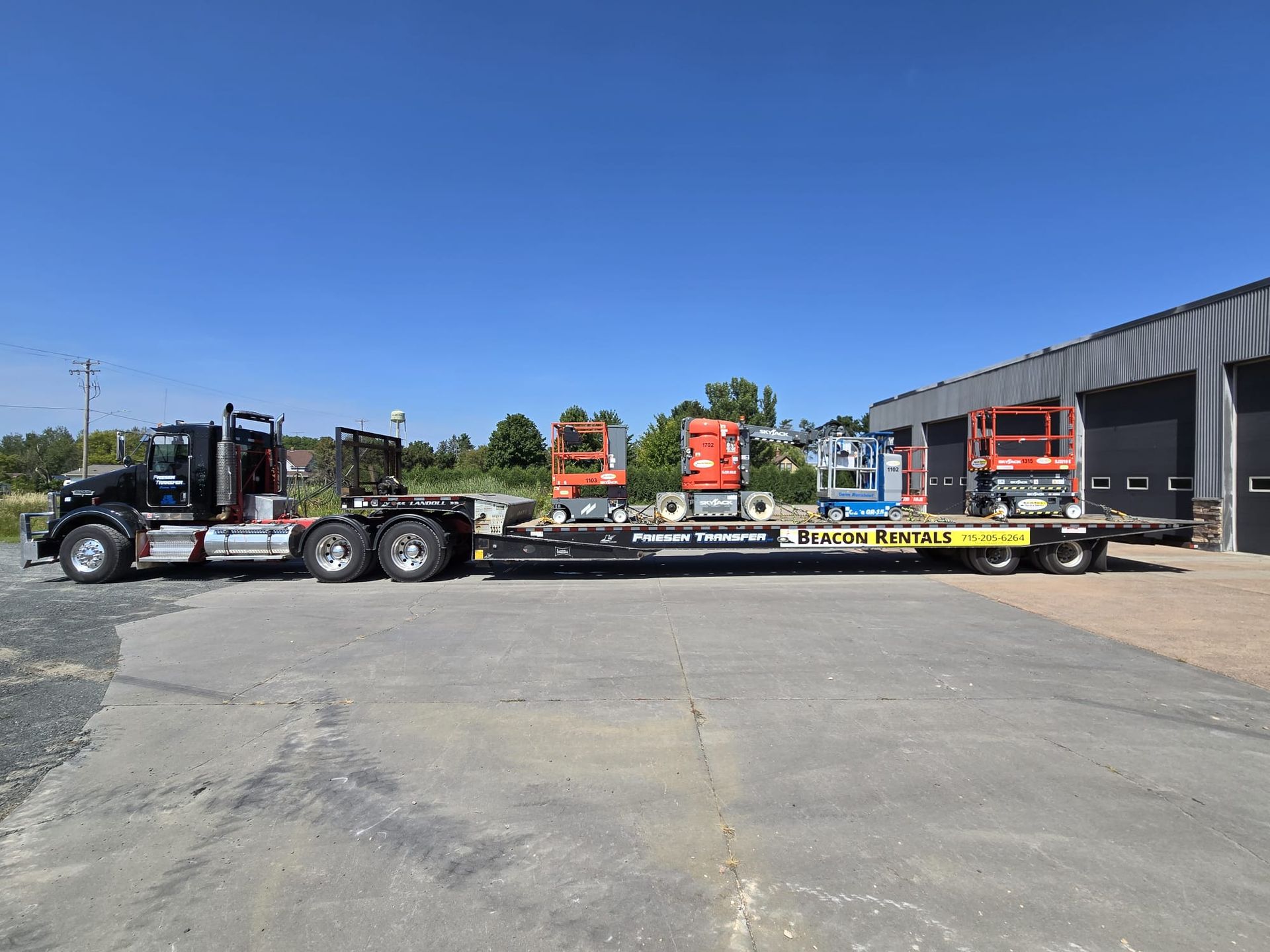 Black truck hauling flatbed trailer with orange and blue aerial lifts, parked outside a building on a sunny day.