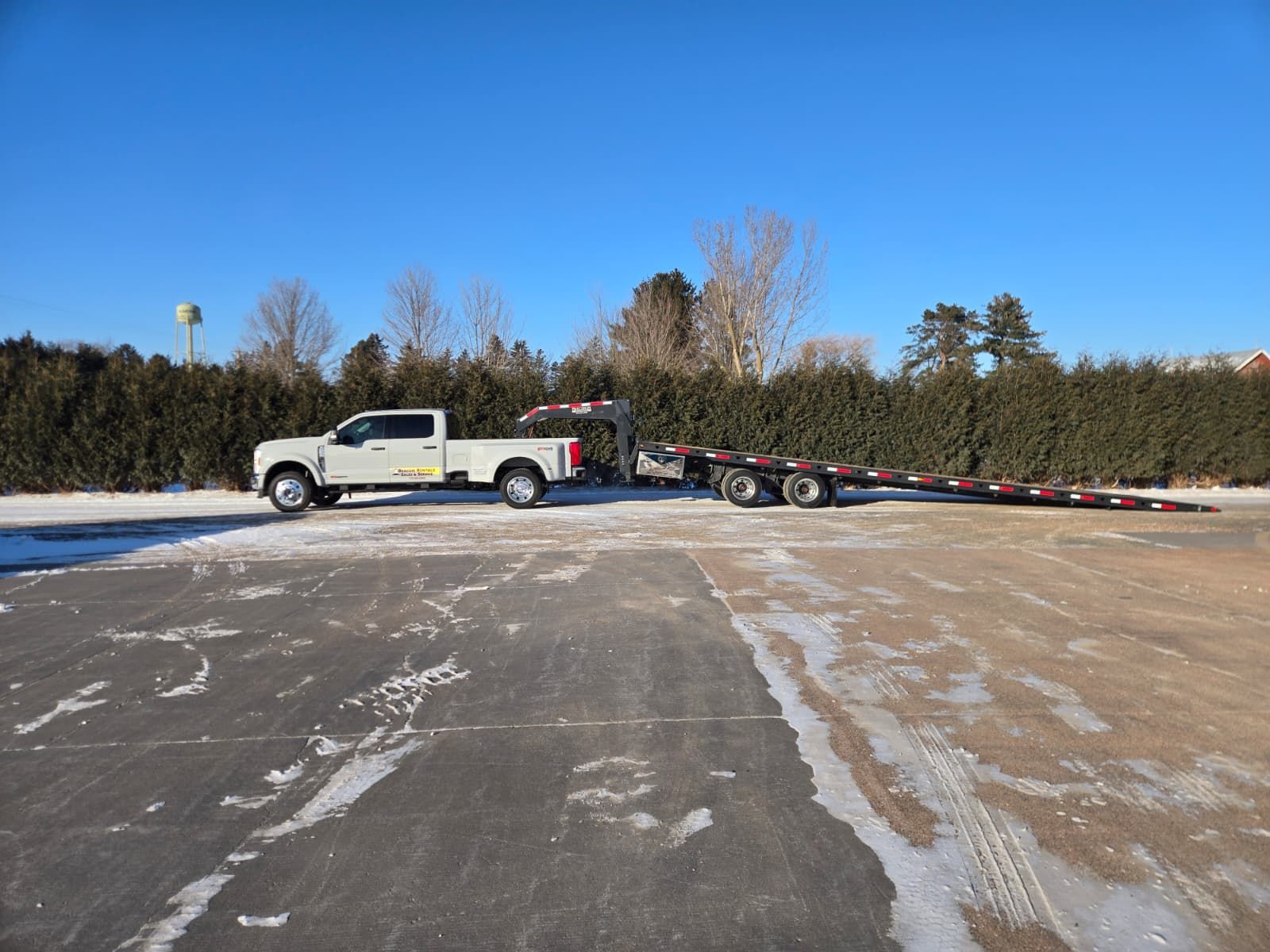 White truck towing a long trailer on a snow-covered parking lot on a sunny day.