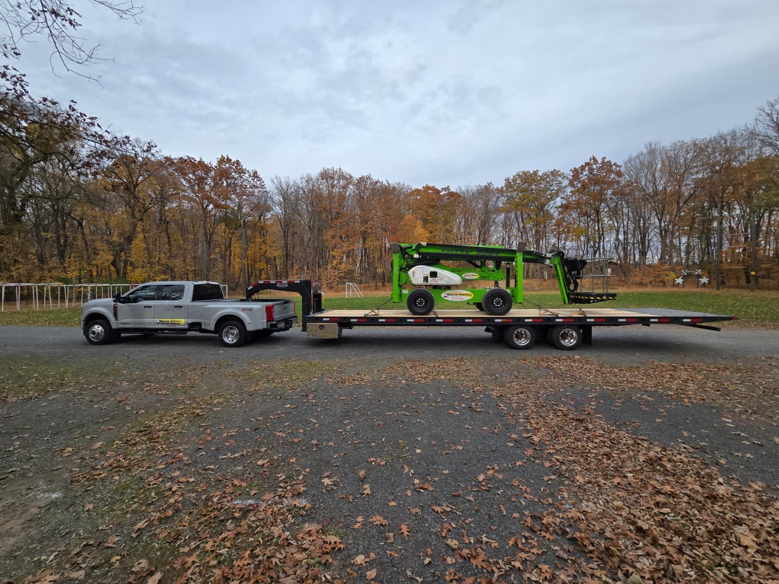 Truck towing a green and black lift on a flatbed trailer in a gravel area with autumn trees.