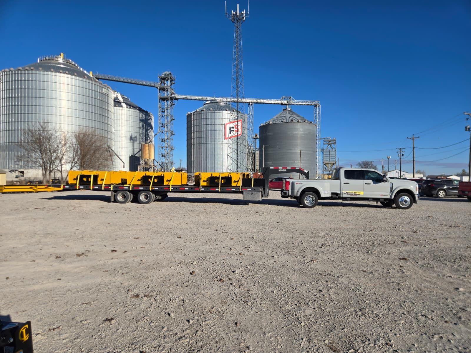 Truck towing a long yellow trailer in front of grain silos under a blue sky.