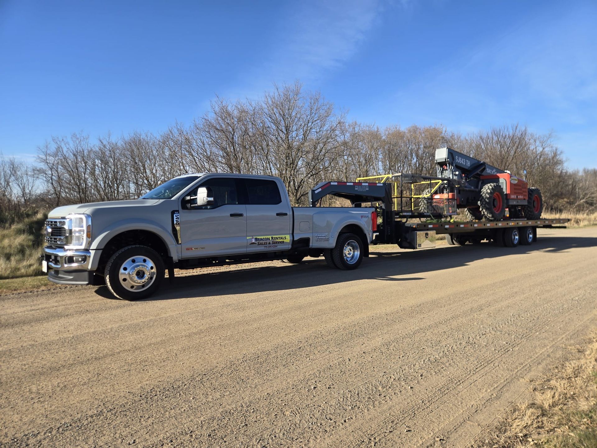 Gray truck towing equipment on a trailer on a dirt road. Trees and blue sky in the background.