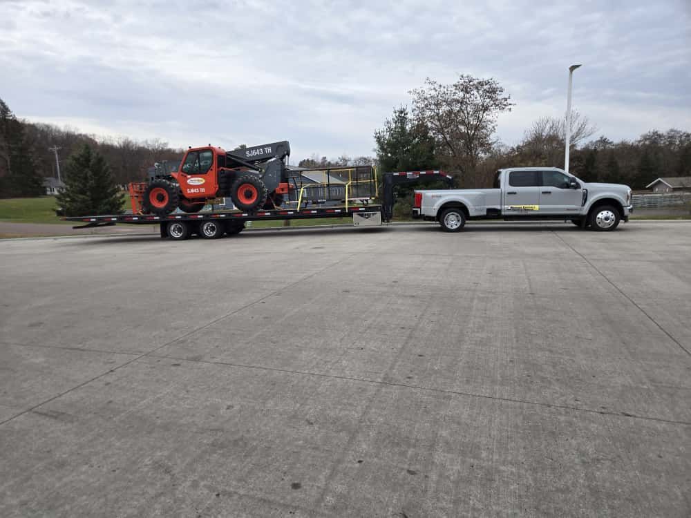 Truck towing a trailer with an orange forklift. The scene is outdoors on a cloudy day.