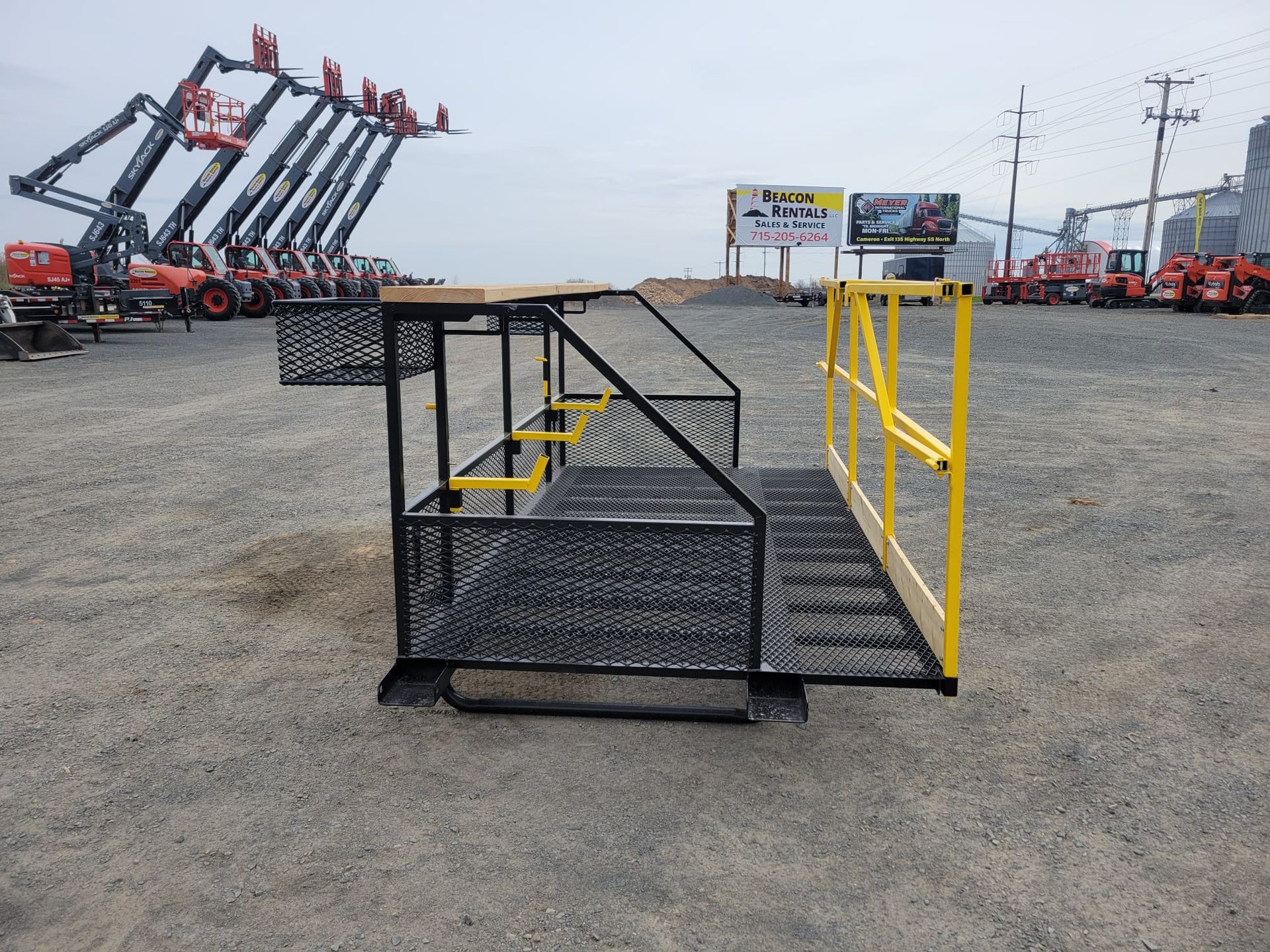 Steel work platform with yellow handrails, black grating, and steps on gravel lot.