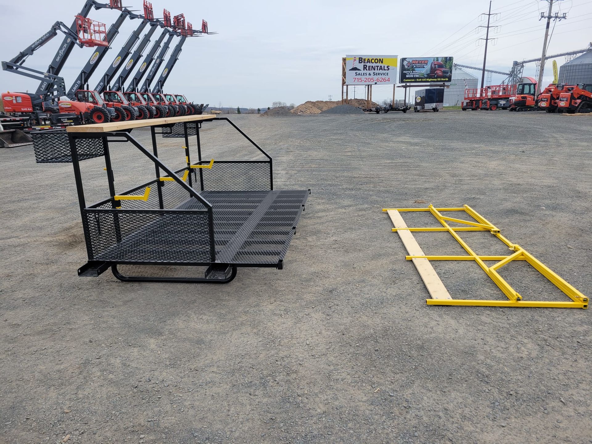 Black metal work platform and a yellow metal frame on a gravel lot; construction equipment in the background.