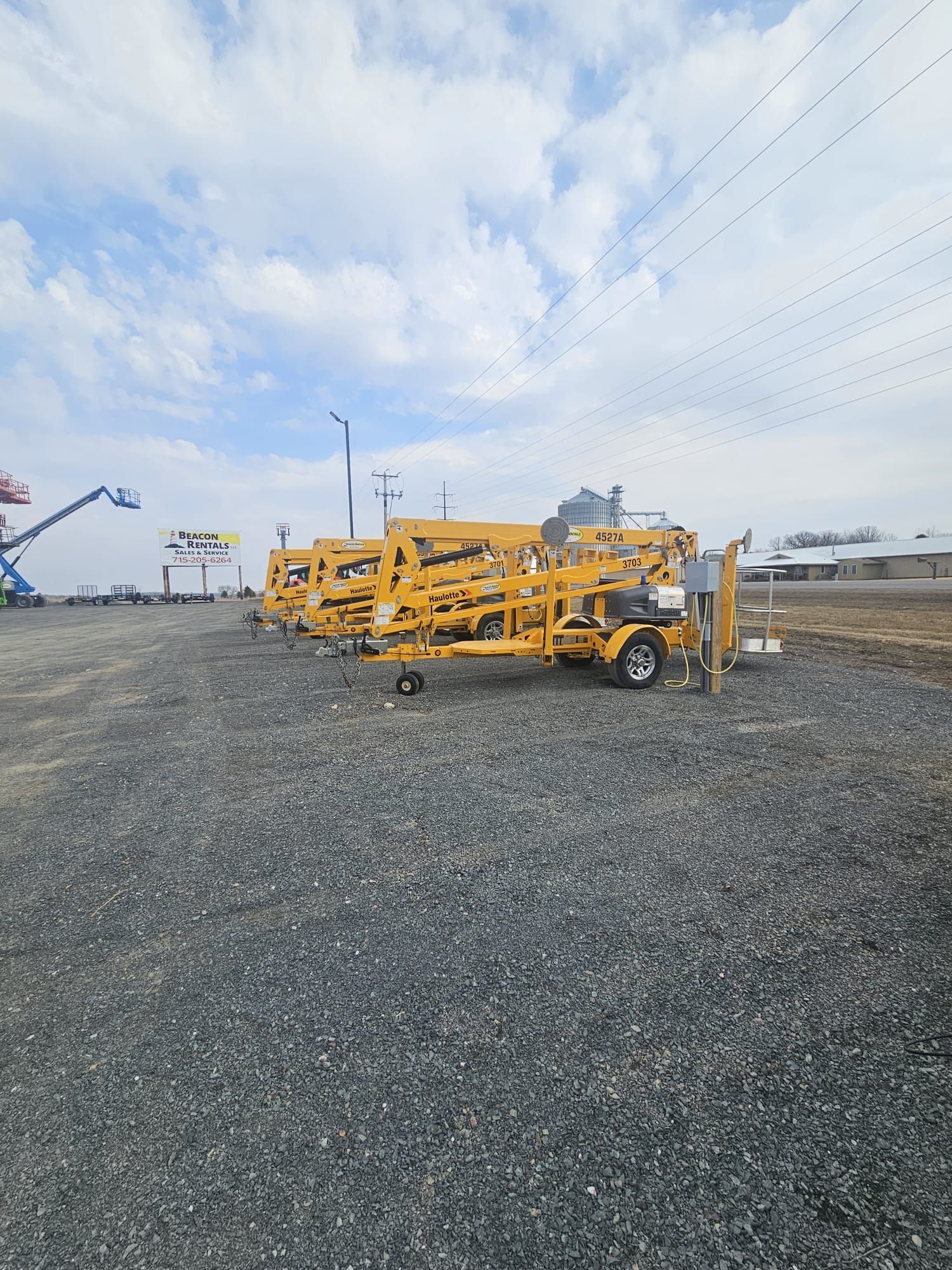 Several yellow boom lifts parked on gravel under a cloudy sky.