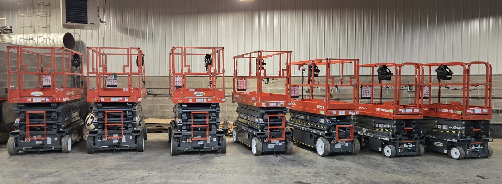 Six red scissor lifts parked inside a warehouse with a corrugated metal wall.