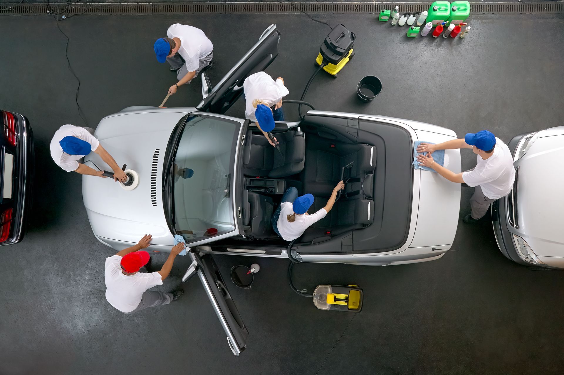Overhead shot: people cleaning a silver convertible car in a car wash.
