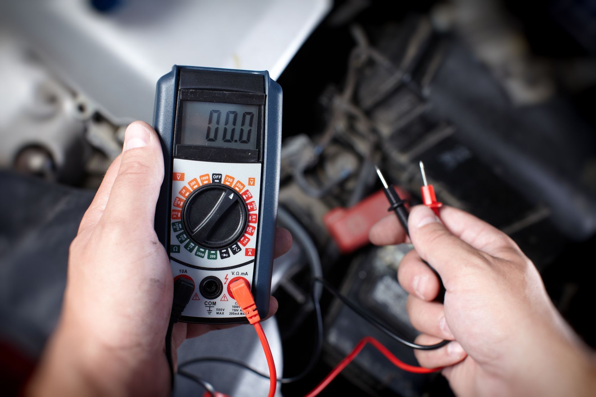 Hands holding a multimeter near a car engine, testing electrical components with the probes.