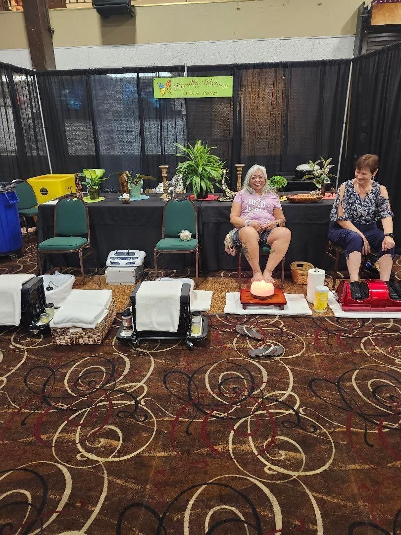 People sitting in massage chairs at an indoor event, with foot spas and a patterned carpet.