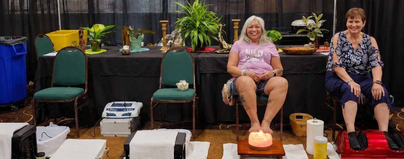 People sitting in massage chairs at an indoor event, with foot spas and a patterned carpet.