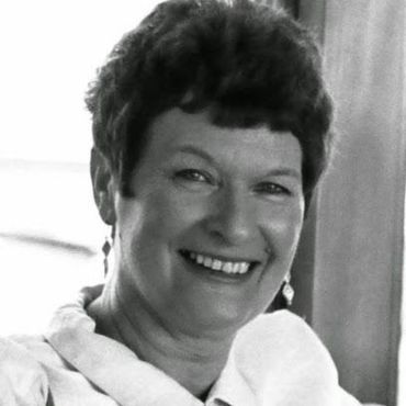 Smiling woman in a white blouse, seated indoors near a window.