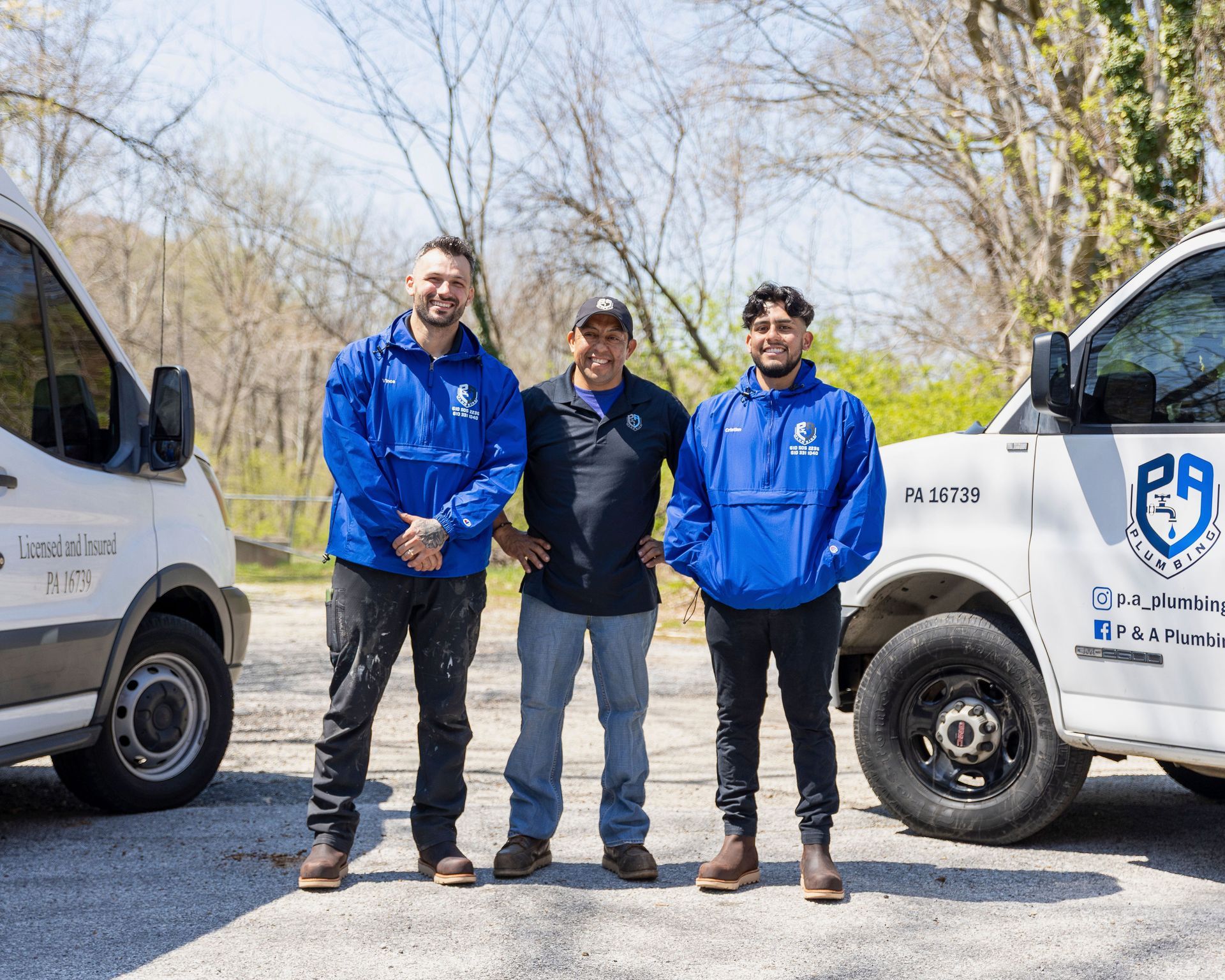 Three workers in blue jackets stand between two white service vans on a sunny road.