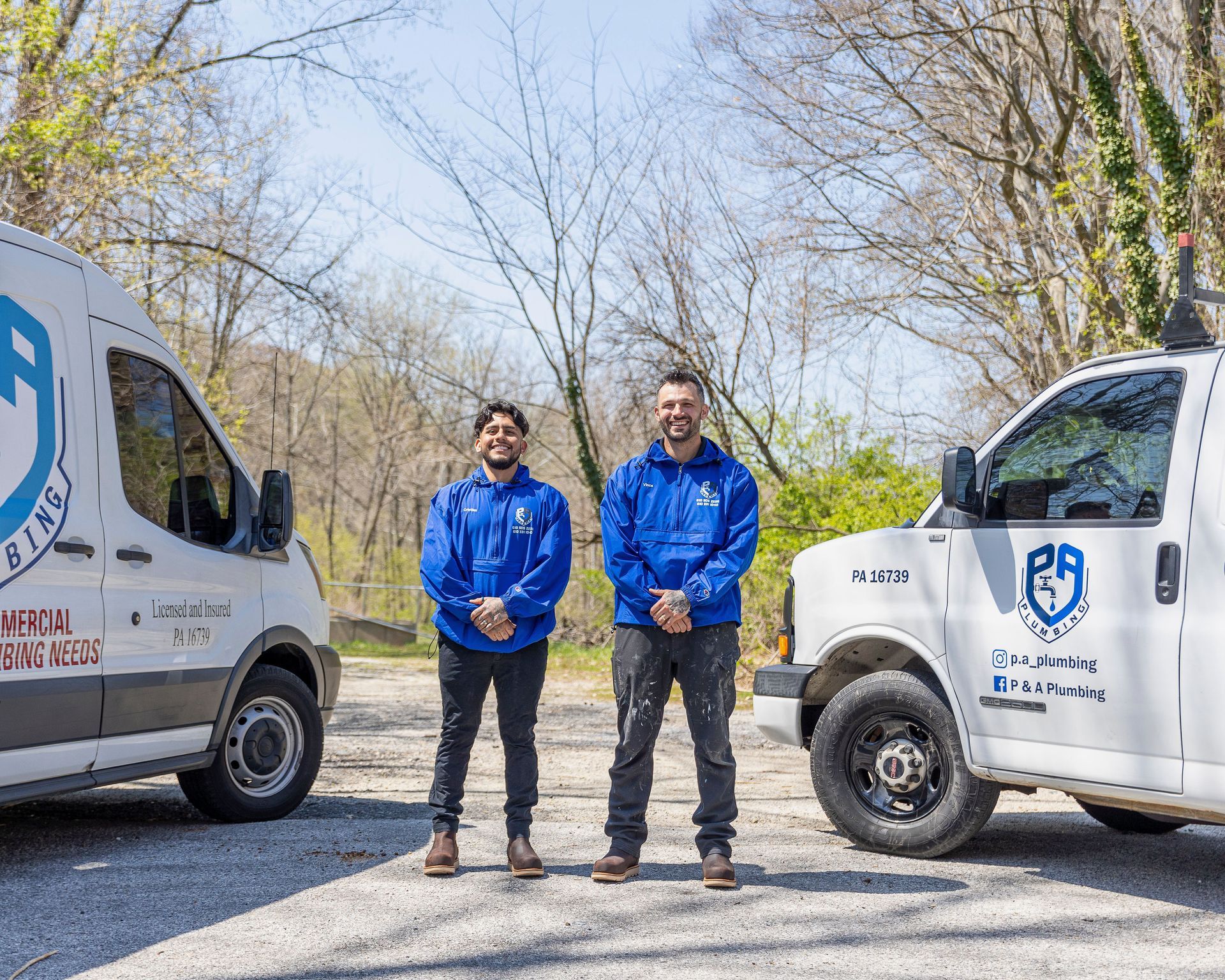 Two workers in blue jackets stand between two white utility trucks on a gravel road