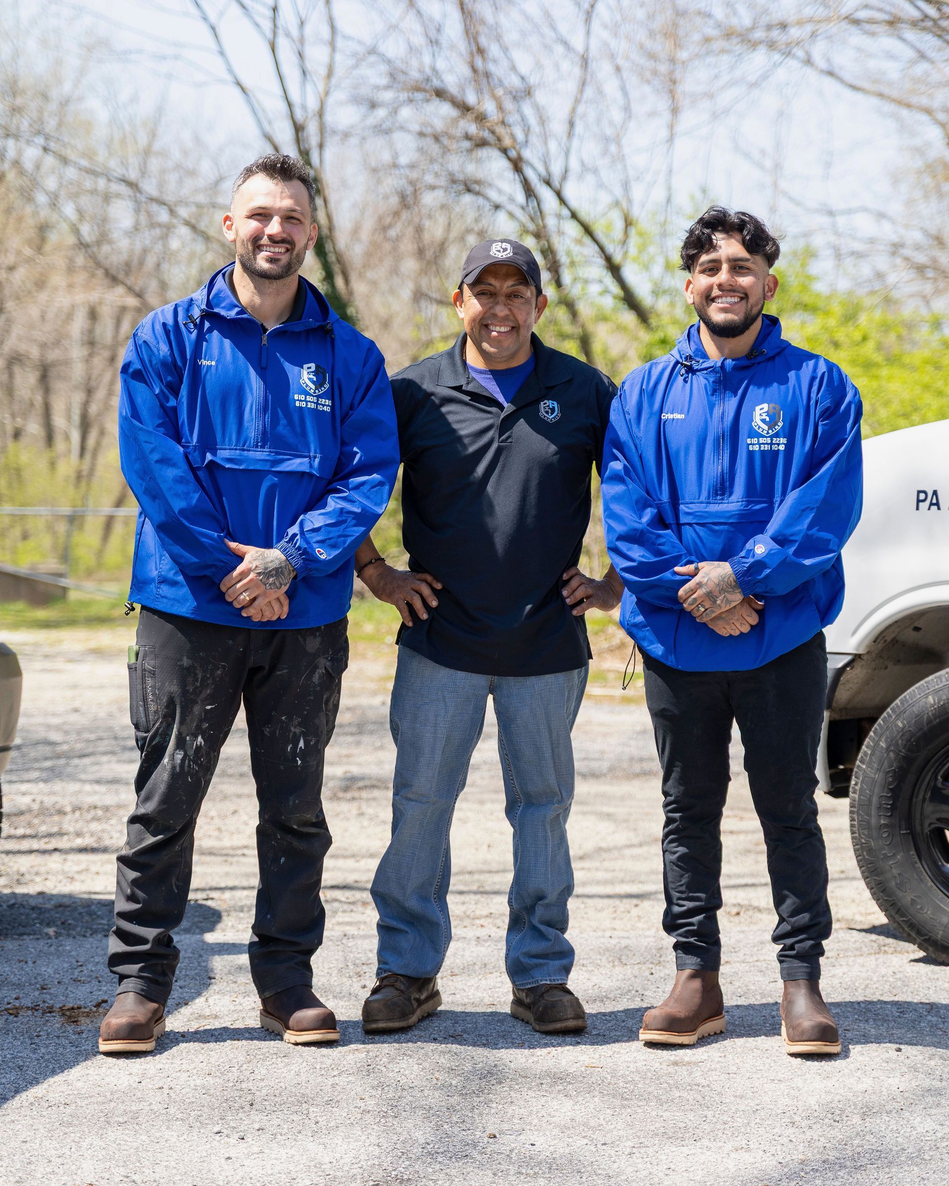 Three people standing outdoors, two in blue jackets, one in a black cap, smiling near a vehicle.