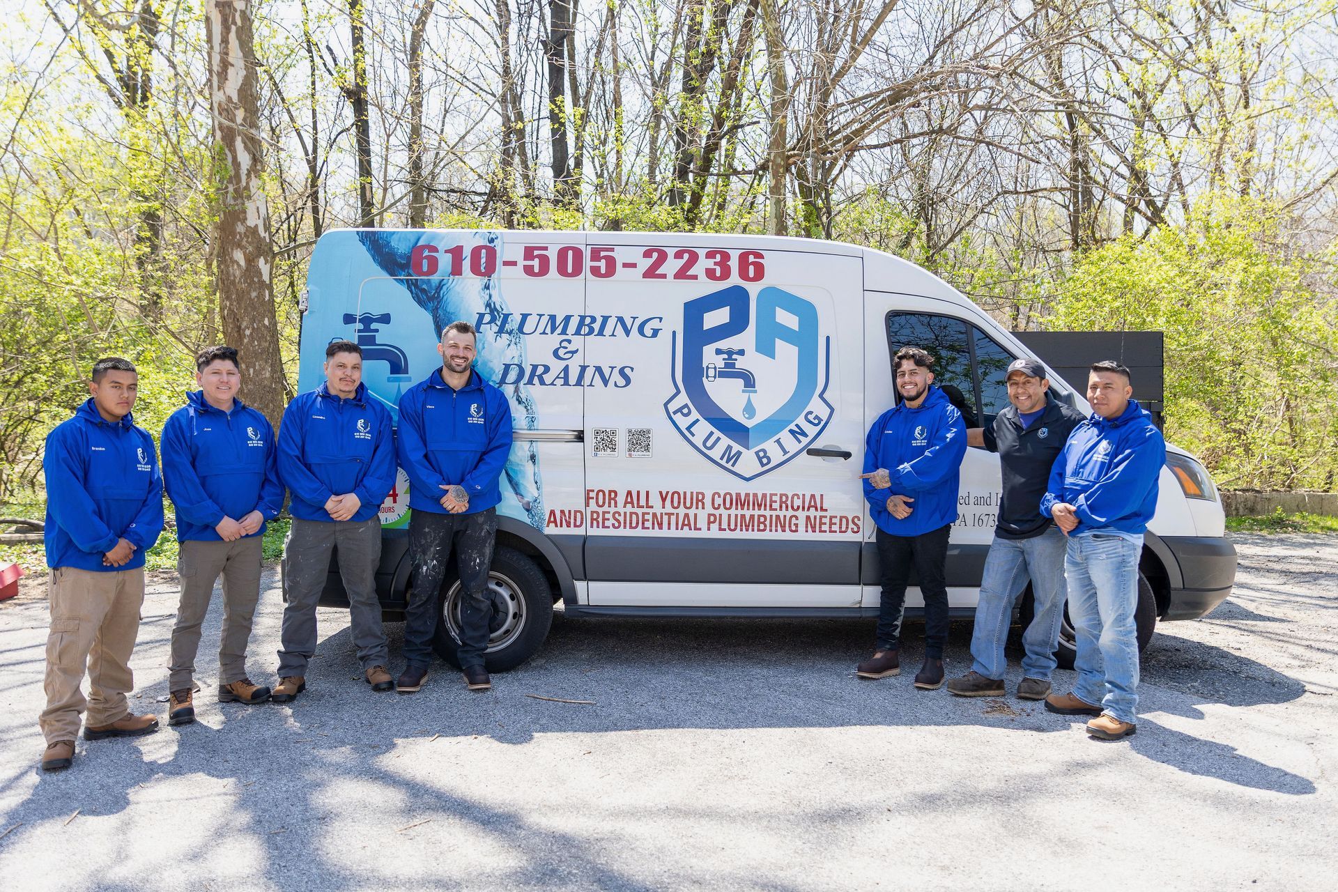 A white service van with blue graphics parked outdoors, with six people in blue jackets standing beside it.
