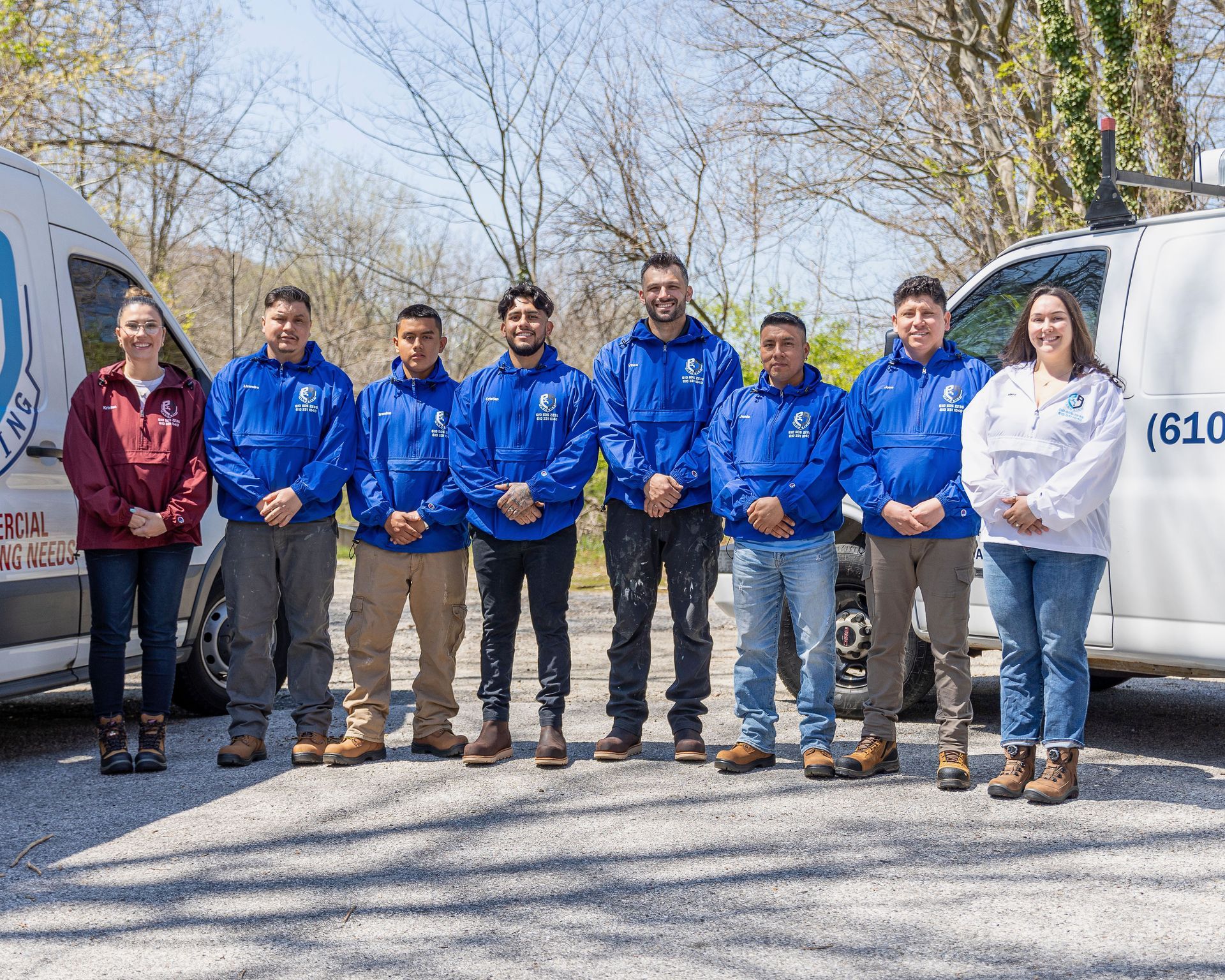 Group of workers in blue jackets standing outdoors between two vans on a sunny day.