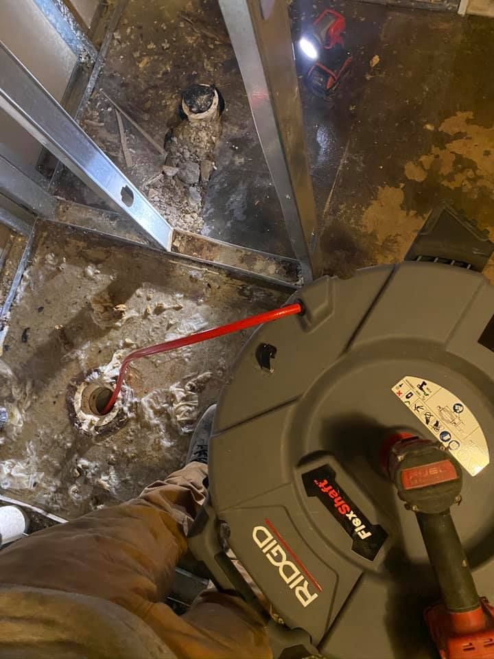 Plumber using a drain snake on a concrete floor near a toilet drain. Gray and orange tools.