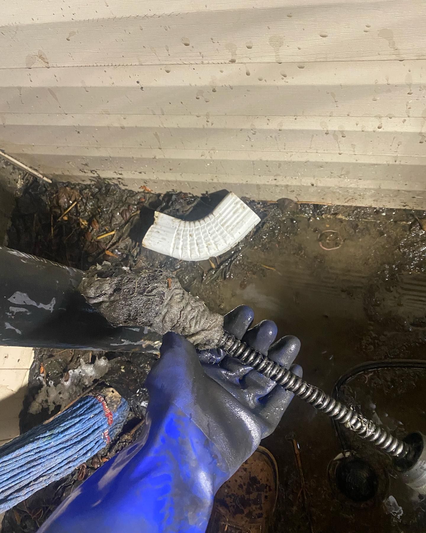 A gloved hand holds a plumbing snake in muddy water, near a white gutter downspout and dirty wall.