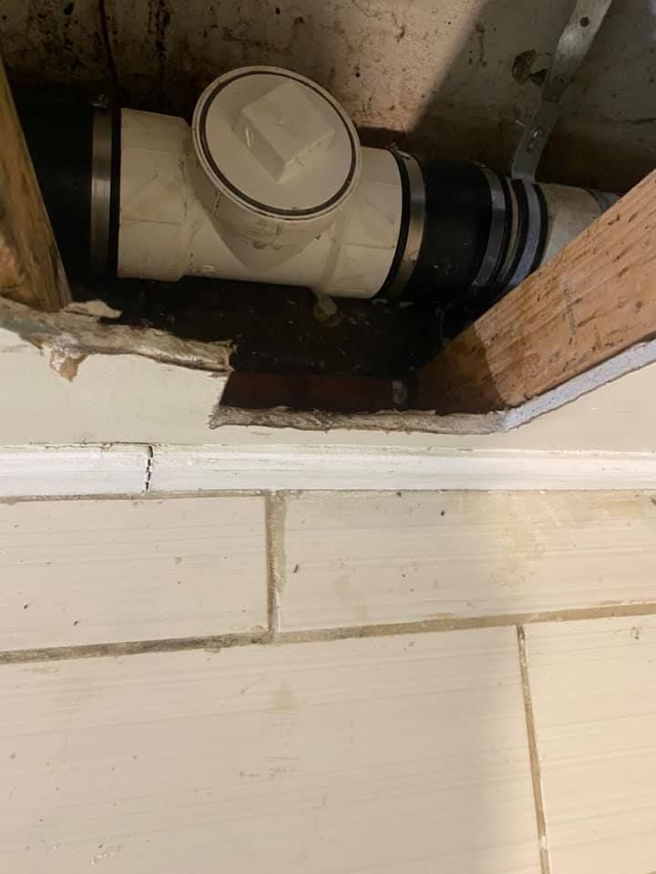 White PVC pipe with cleanout cap running through a rough wall opening above a tiled wall.
