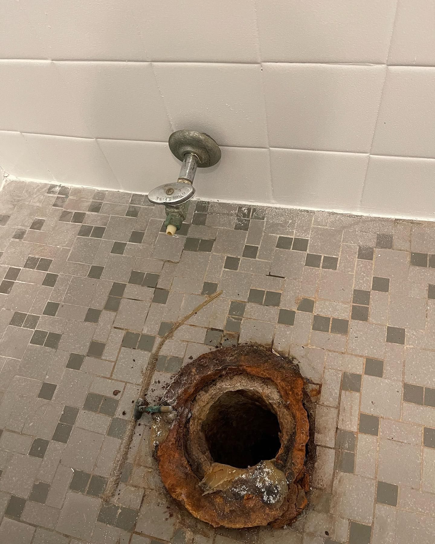 Rusty toilet drain hole in a tiled bathroom floor beneath a wall-mounted pipe.