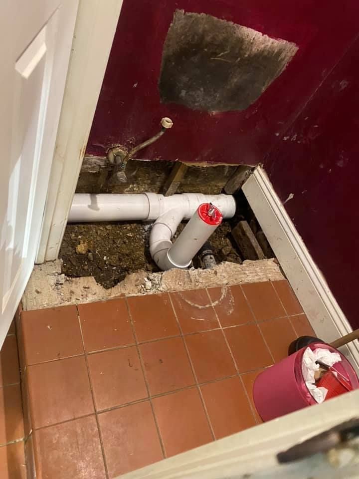 Bathroom floor cut open around exposed pipes, with broken tile, dirt, and a wall patch visible.