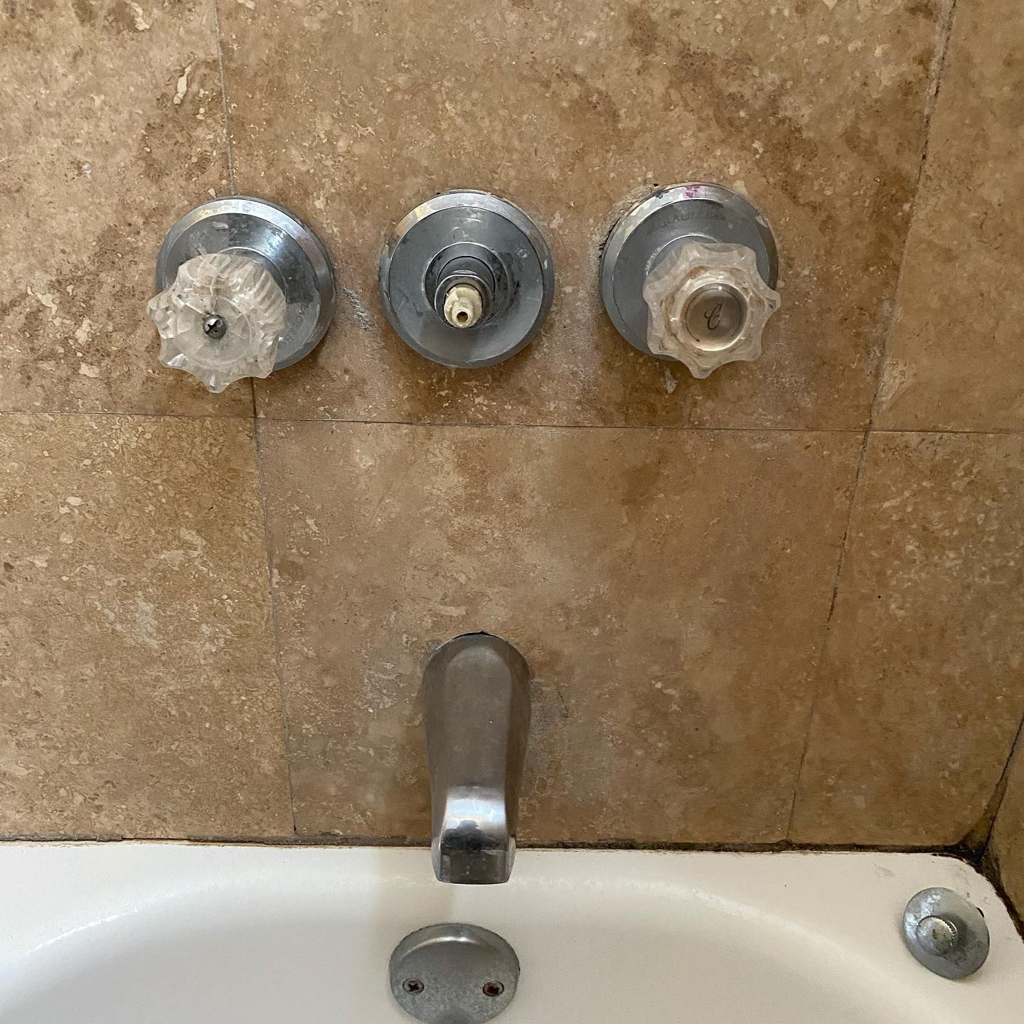 Bathtub faucet with three valve knobs, and a white tub set against beige tile.