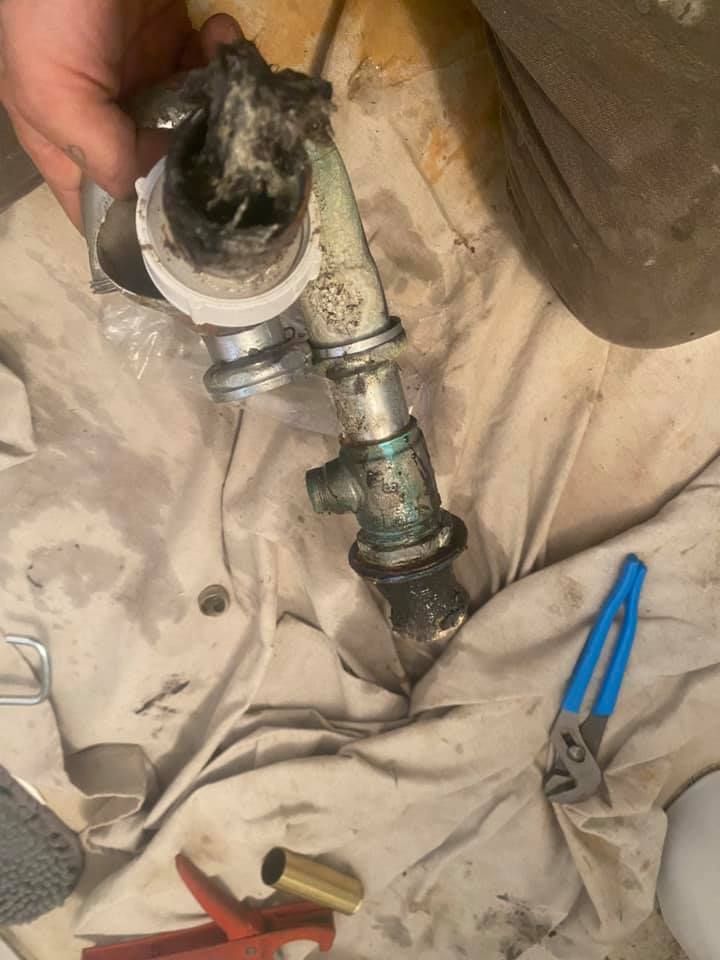 Hand holding a pipe fitting over a beige tarp, with a metal pipe and blue pliers nearby.