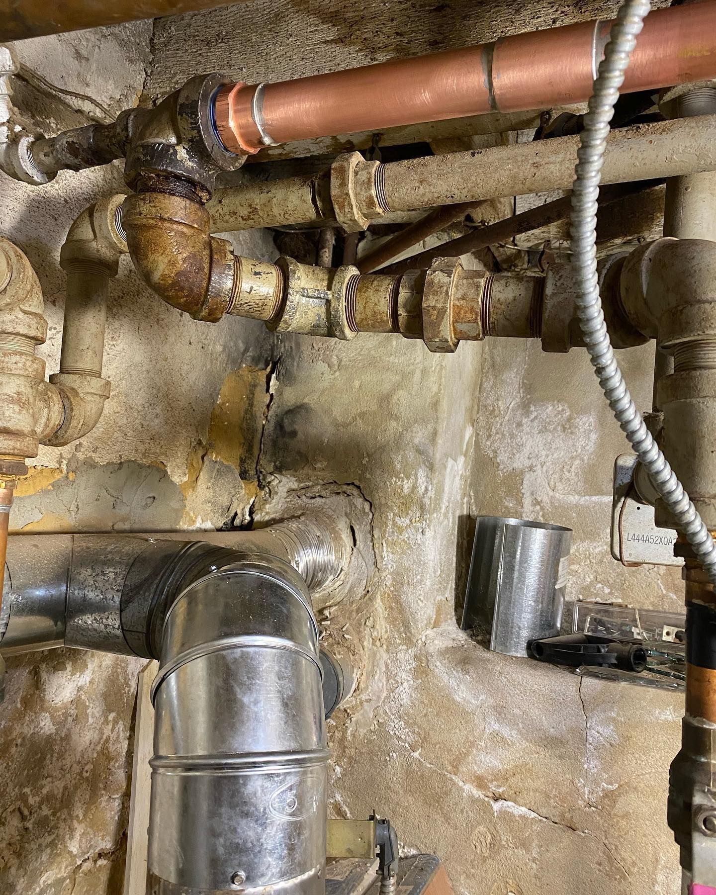 Rusty basement pipes and fittings on a stained concrete wall with a silver vent pipe.