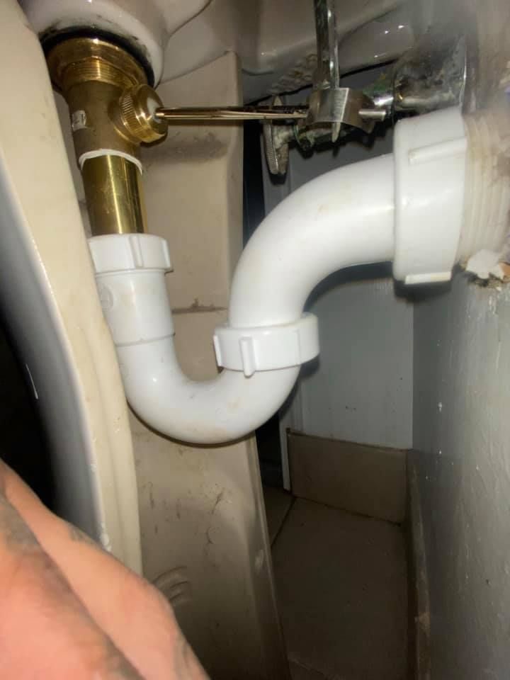 Under-sink plumbing with white PVC drain pipes connected to a brass pipe under a cabinet.