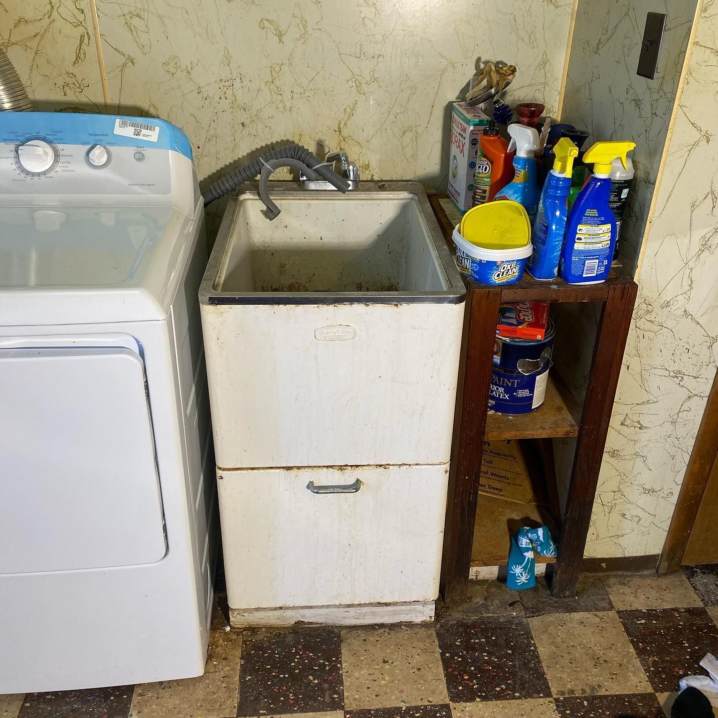 A laundry room with a white sink, dryer, and shelves holding cleaning supplies.