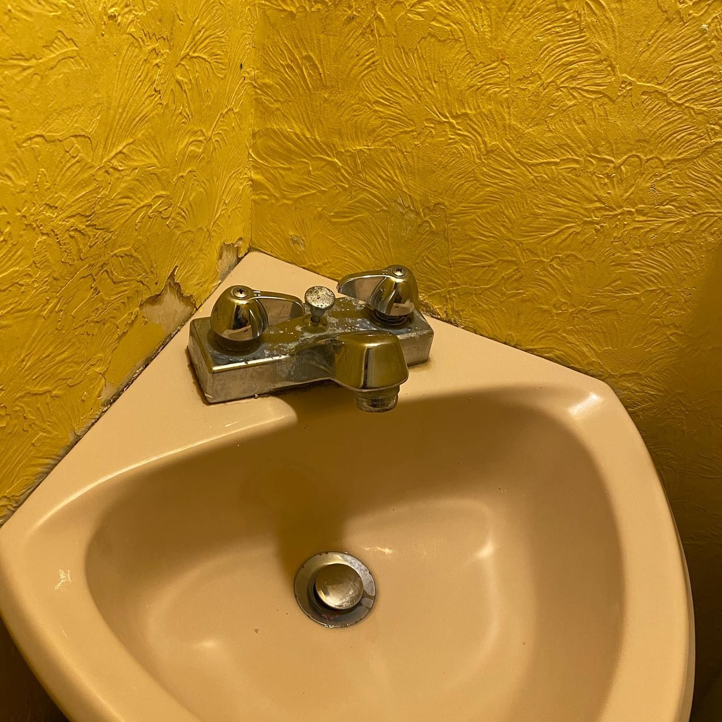 Corner sink, cream-colored, with chrome faucet, against a yellow textured wall.