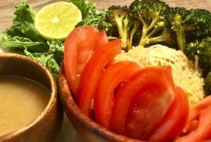 Fresh vegetables, lemon, and dipping sauce arranged on a plate with a noodle dish in the background