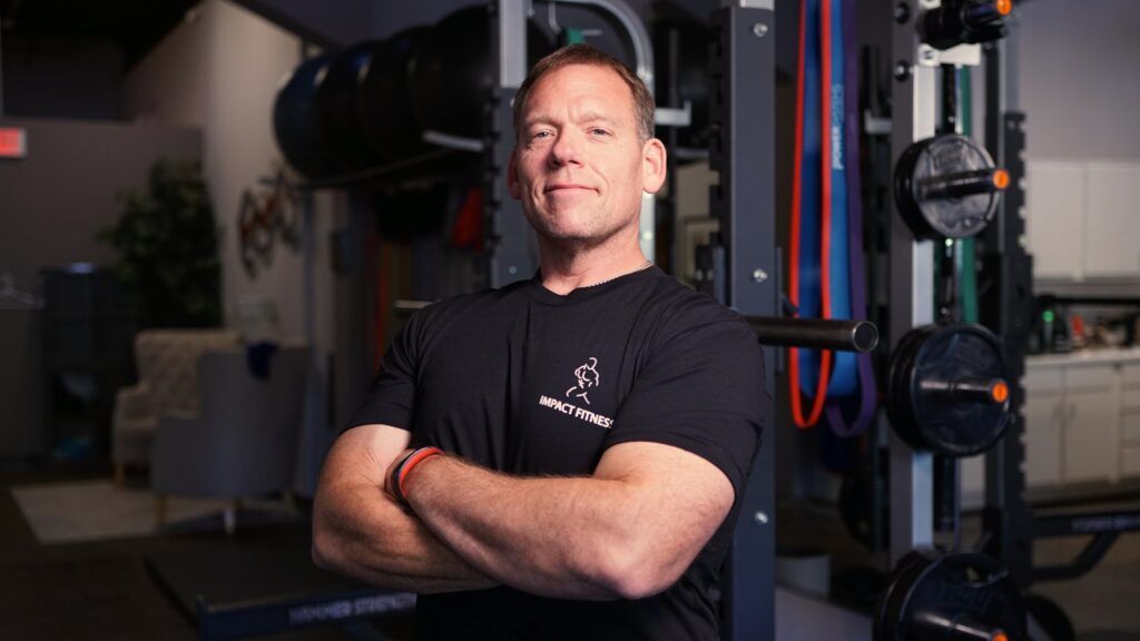 Man with folded arms standing in a gym beside weight equipment, wearing a black T-shirt.