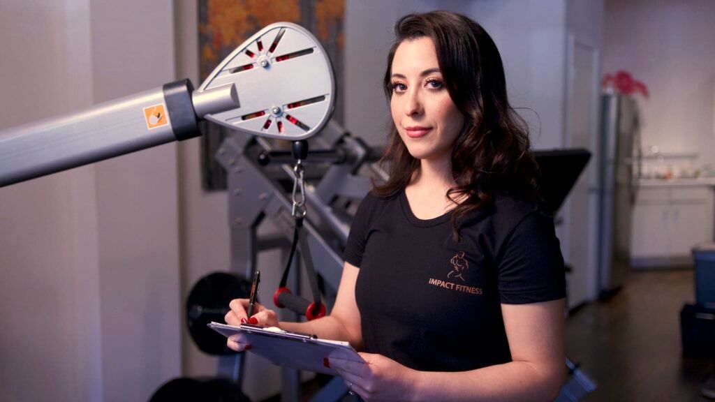 Woman in a lab, holding a device beside a mounted radar dish in an industrial setting.