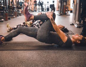 Woman stretching on gym floor with one leg crossed over her chest, exercise equipment in background