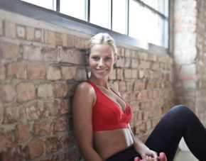 Woman in a red sports bra seated by a brick wall, smiling in a loft-style gym setting.