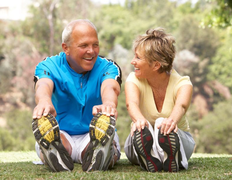 Two people in workout clothes stretching on grass outdoors, smiling at each other.