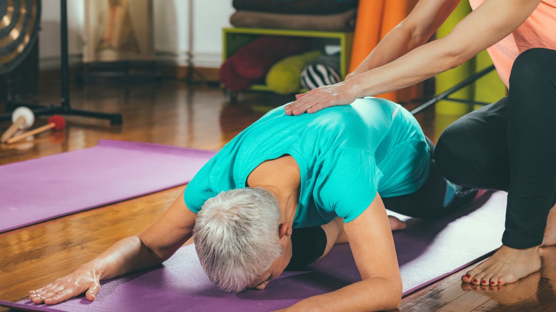 Older person doing yoga stretch on a mat while another person assists in a bright studio