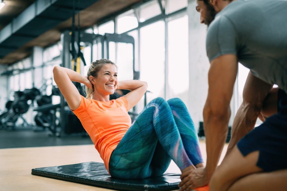 Woman doing crunches on a mat in a gym, with a trainer nearby.