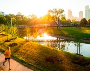 Sunlit park with a runner on a path beside a river and bridge, city skyline in the distance