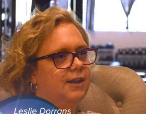 Woman with short blonde hair and glasses speaking indoors, seated on a beige couch.