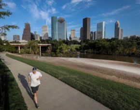 Runner on a paved trail beside a grassy riverbank, with a city skyline in the background.