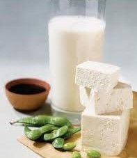 Tofu blocks, green edamame pods, a bowl of soy sauce, and a glass of soy milk on a wooden board