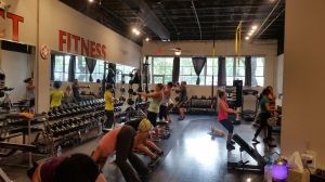 People working out in a bright fitness studio with mirrors, dumbbells, and exercise mats.